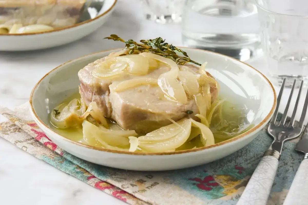 Un trozo de atún en escabeche con cebolla y hierbas, servido en un plato blanco con un tenedor al lado.