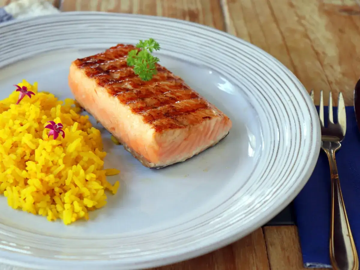 Delicioso salm&oacute;n a la plancha con marcas de parrilla, servido con arroz amarillo y decorado con perejil y flores.