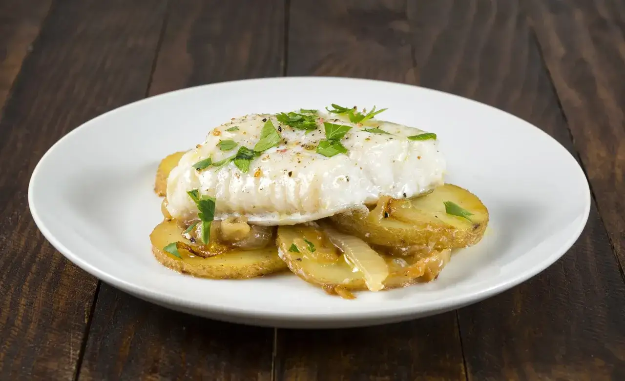 Filete de bacalao sobre patatas asadas y cebolla caramelizada, perfecto para una receta bacalao al horno sencilla. Decorado con perejil fresco.