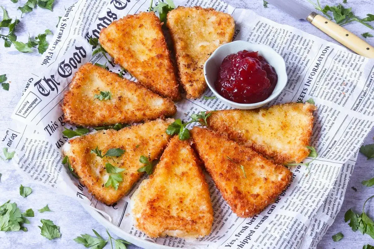 Tri&aacute;ngulos dorados de queso frito con mermelada roja en un plato con peri&oacute;dico y perejil.
