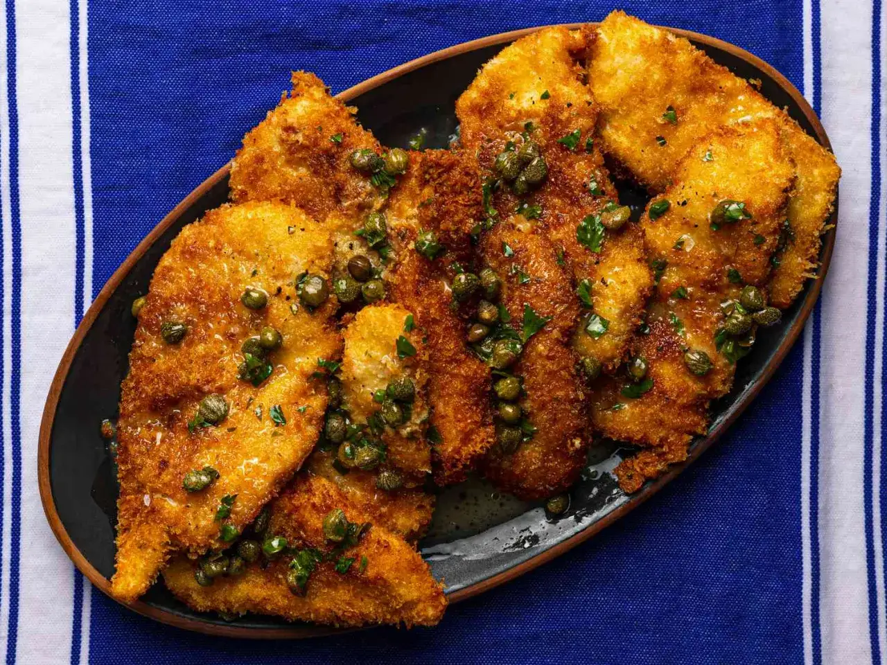 Un delicioso escalope de pollo empanizado, bañado en una salsa dorada con alcaparras y perejil fresco, servido en un plato oscuro sobre un mantel azul a rayas.
