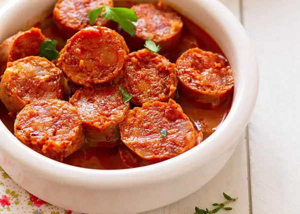 Chorizos a la sidra jugosos y dorados en una salsa rojiza, servidos en un cuenco blanco.