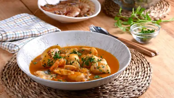 Deliciosa cazuela de pescado con gambas y trozos de pescado blanco en una salsa rojiza, adornada con perejil fresco.