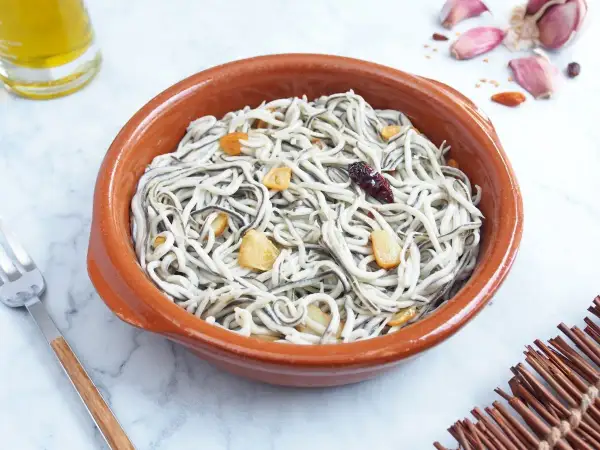Un cuenco de barro con gulas al ajillo, salteadas con ajo y guindilla. Se ve un tenedor y una botella de aceite de oliva.