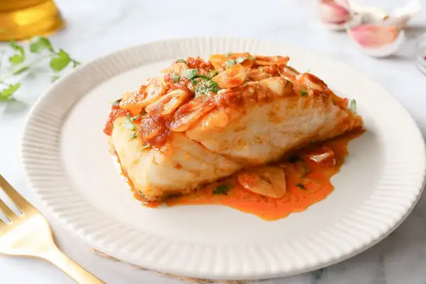Bacalao a la llauna jugoso, cubierto de salsa de tomate y ajo laminado, servido en plato blanco con tenedor dorado.