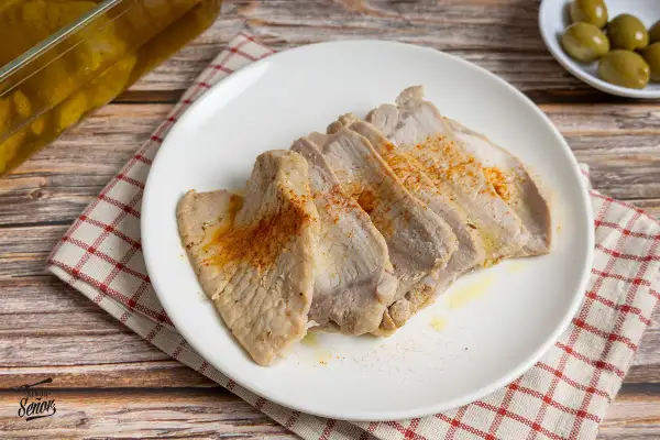 Lomo de orza tierno y jugoso, cortado en lonchas y espolvoreado con pimentón. Servido en plato blanco sobre mantel a cuadros.