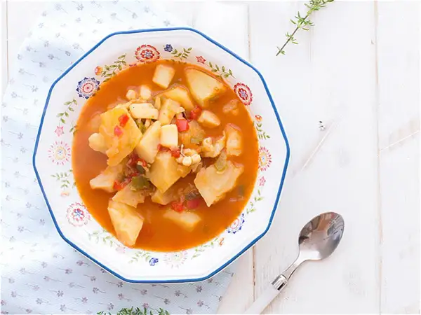 Un plato de sepia con patatas, pimientos y trozos de tomate en una salsa rojiza, servido en un cuenco decorado.