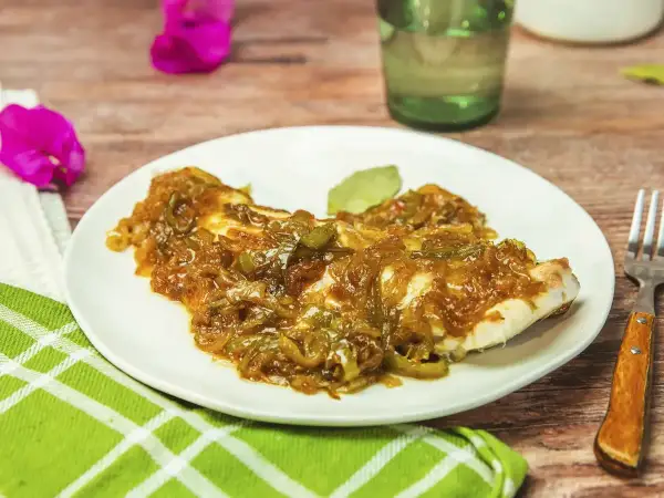 Un plato de urta a la roteña, con pescado blanco cubierto de cebolla y pimientos pochados, servido en una mesa de madera.