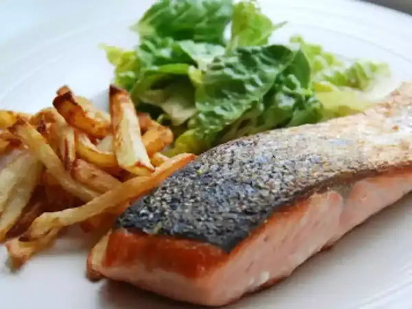 Un delicioso salmón a la plancha con piel crujiente, acompañado de patatas fritas doradas y una fresca ensalada verde.