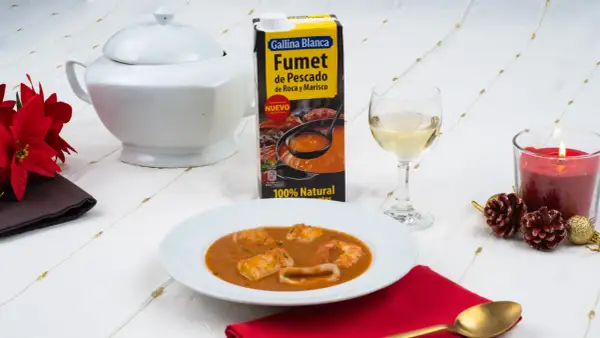 Caldereta de pescado servida en plato blanco, acompañada de fumet de pescado y marisco, vino blanco y vela encendida.
