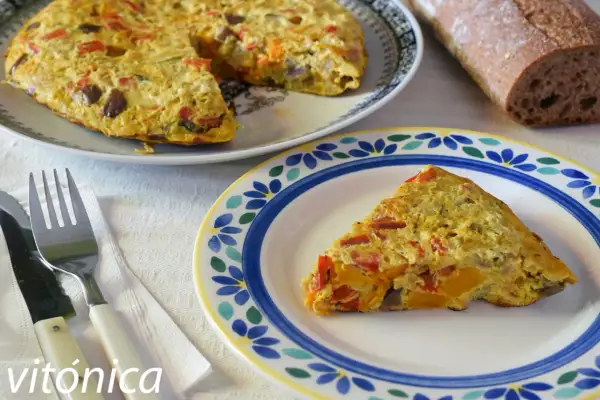 Deliciosa tortilla de berenjena con pimientos y calabaza, servida en un plato decorado. Acompañada de pan rústico y cubiertos.