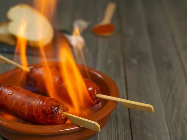 Chorizo al infierno, dos salchichas en brochetas ardiendo en una cazuela de barro, con pan y pimentón al fondo.